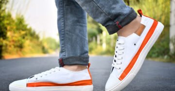 A pair of white and orange men's sneakers styled with rolled-up denim jeans, displayed on a road with a blurred natural background.